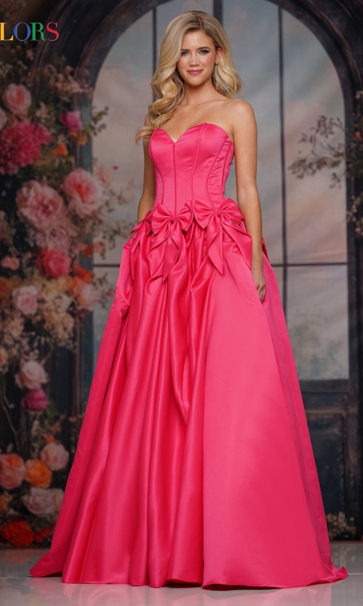 A woman stands before a floral and window backdrop, wearing the Bow-Front Long Prom Ball Gown (Colors Dress 3739) in hot pink, featuring a strapless sweetheart neckline, fitted bodice, and decorative bows.